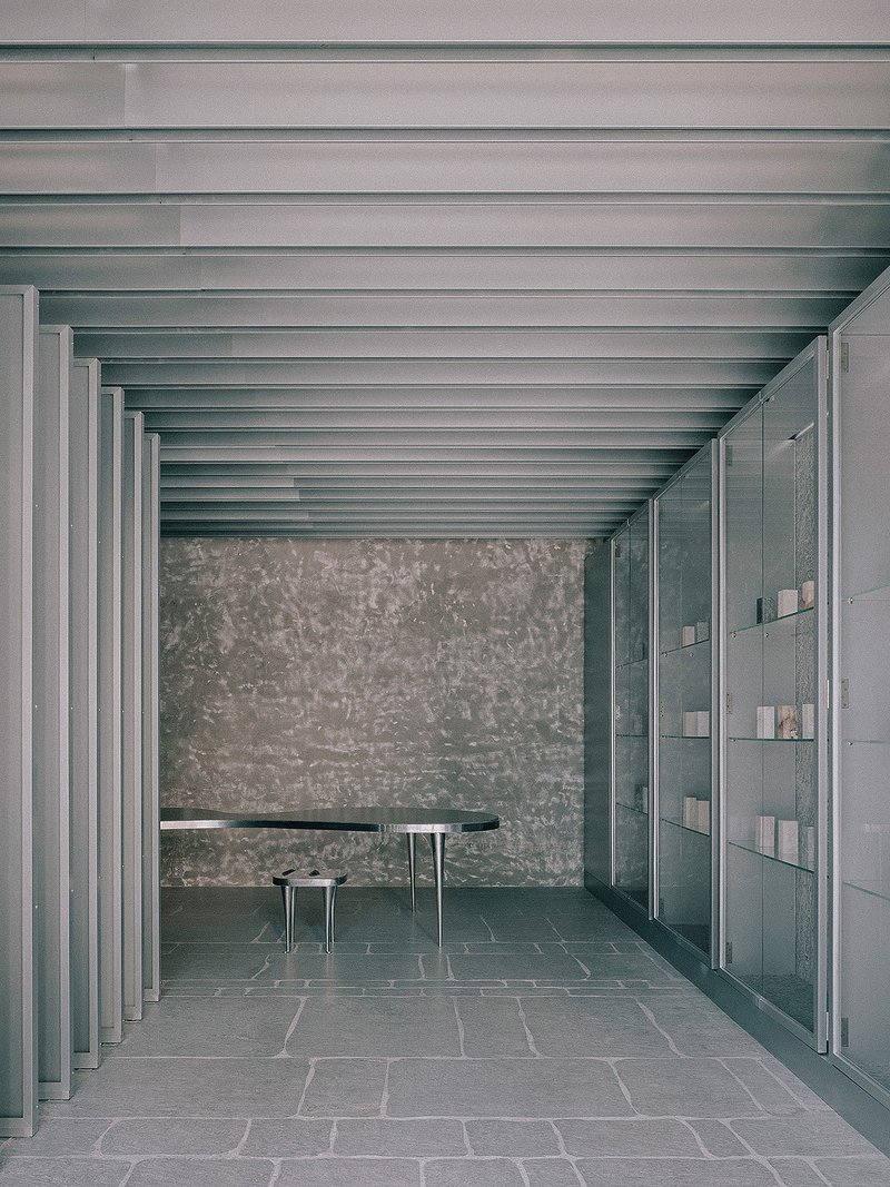 Interior colonnade with exposed metal beams, textured stone wall, dark table and floor-to-ceiling glazed partition