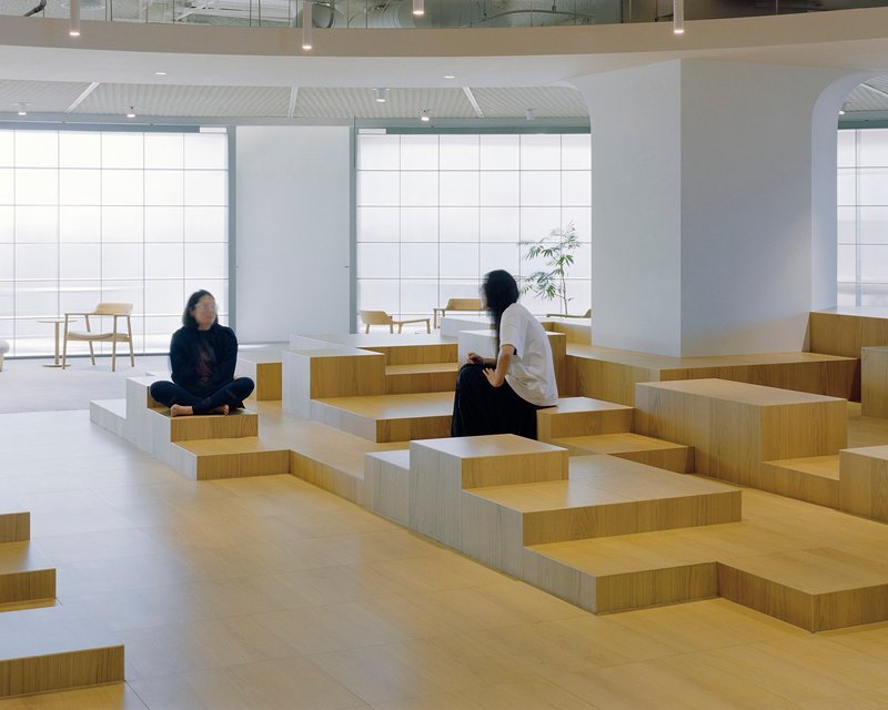 Two visitors seated on geometric timber block seating near gridded translucent glass facade