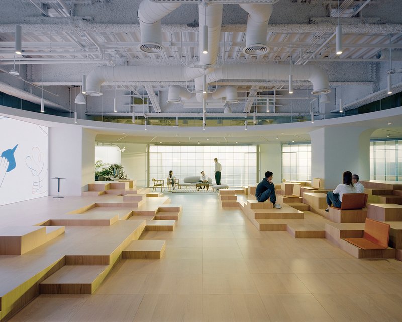Tiered plywood seating platforms in an open office atrium with exposed ductwork and mezzanine above