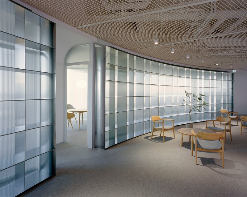 Interior lounge with curved translucent glass panel wall and expanded metal ceiling under recessed lighting