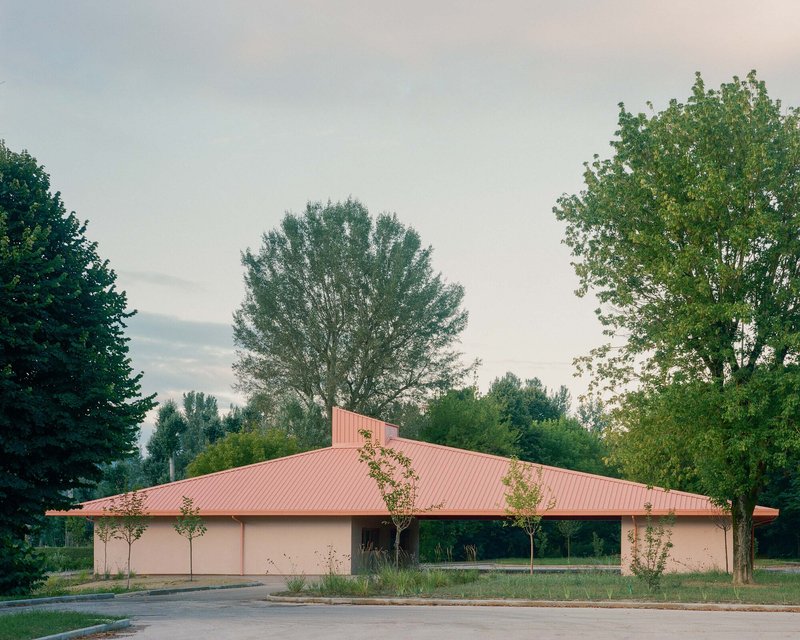 Single-storey structure with pink corrugated metal roof and stucco walls framed by tall deciduous trees