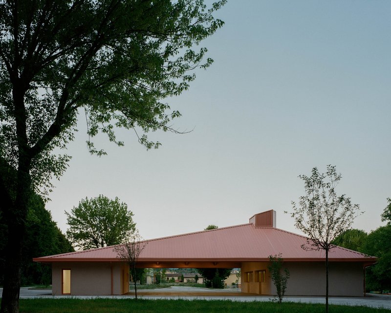 Pavilion with broad hipped metal roof and open-air loggia framed by young trees at dusk