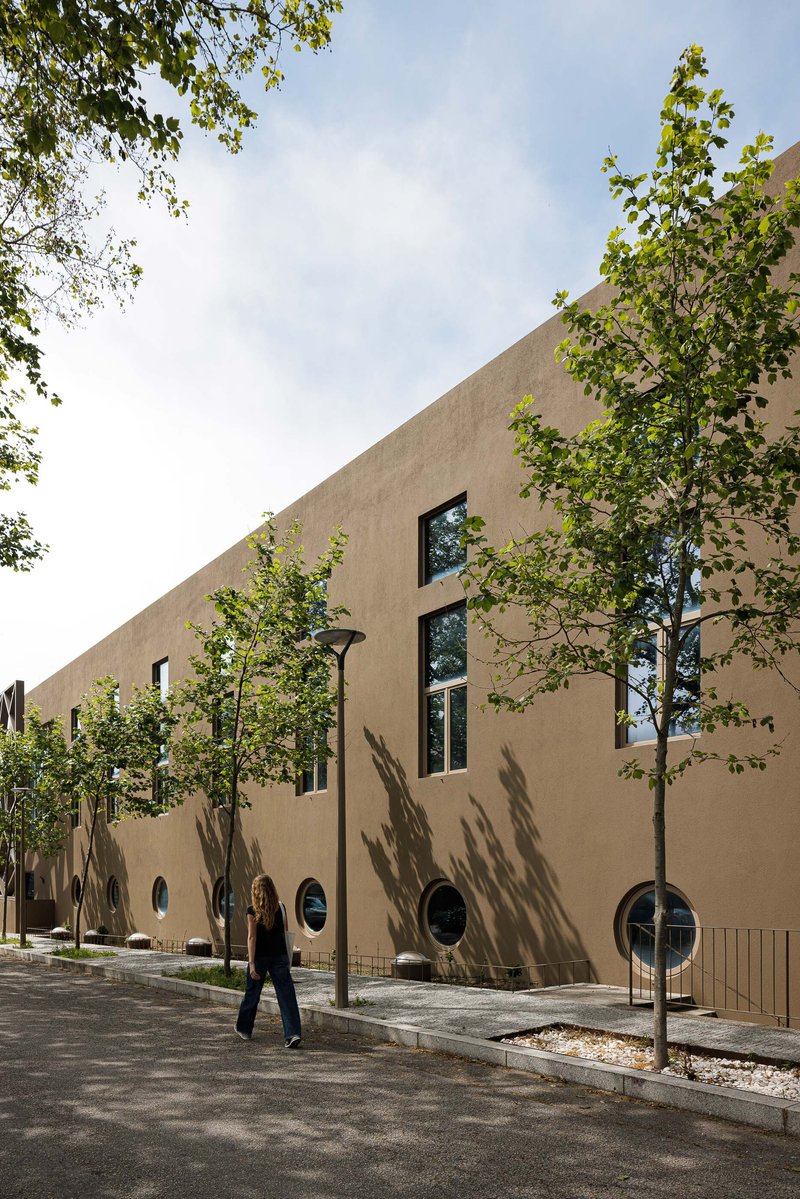 Street-facing facade with circular and rectangular windows and young trees casting shadows on beige stucco