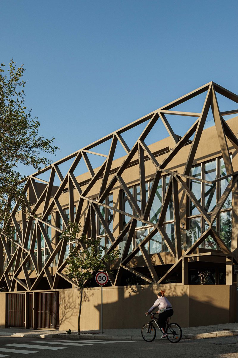 Diagonal steel lattice wrapping the upper volume as a cyclist passes beneath a clear blue sky