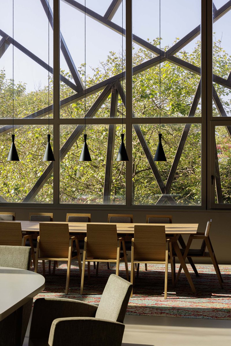Interior view through floor-to-ceiling glazing framed by diagonal steel members overlooking a dining area and trees
