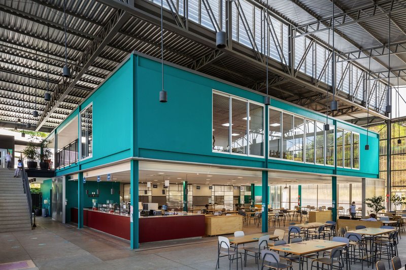 Turquoise two-story volume with cafeteria and dining tables beneath an exposed corrugated metal roof structure
