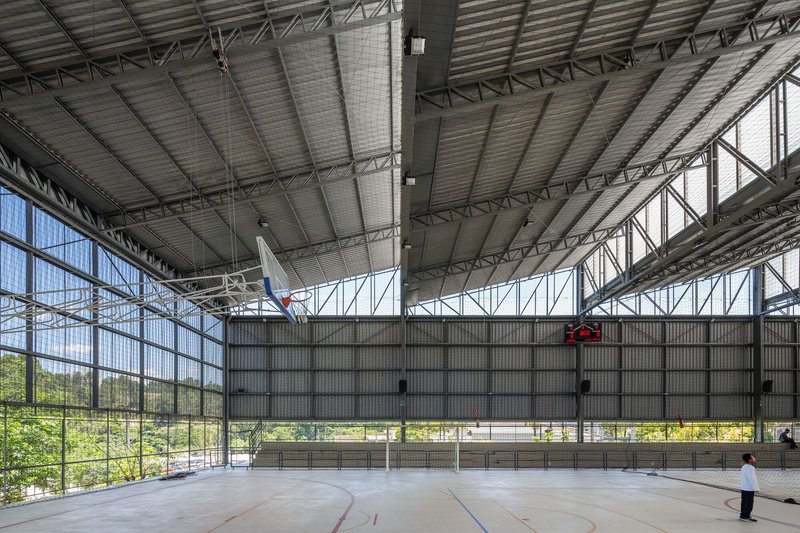 Interior sports court with corrugated metal ceiling and exposed steel trusses overlooking greenery through glazed walls