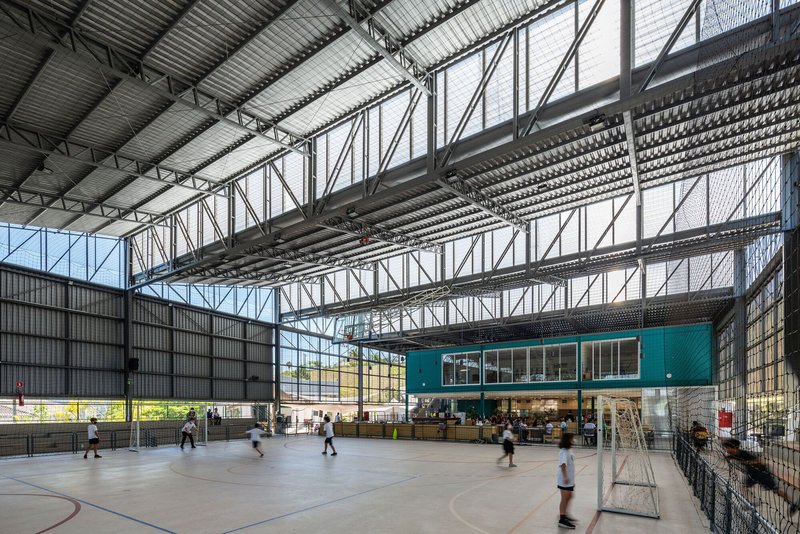 Basketball court with players in motion beneath diagonal metal mesh and glazed wall showing trees outside