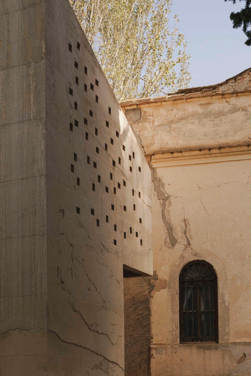 Perforated concrete wall rising beside weathered masonry facade with arched door and tree beyond