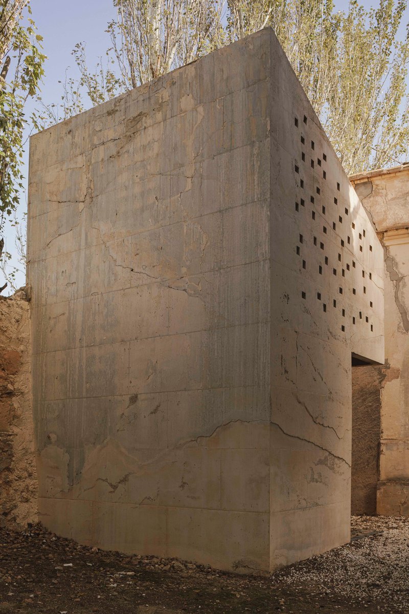 Monolithic concrete volume with irregular perforations standing on gravel beside a historic wall