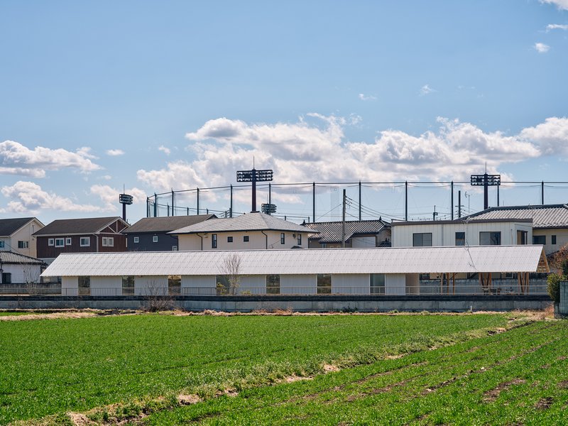 Low horizontal volume with white corrugated metal roof beyond green agricultural field and suburban houses