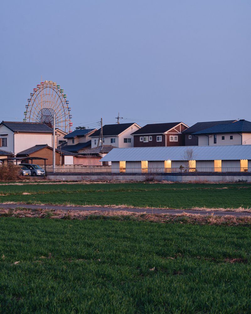 Long facade with evenly spaced illuminated openings viewed across a field at dusk with a ferris wheel behind