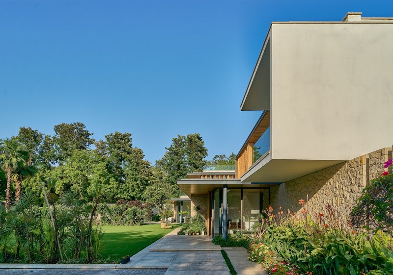 Cantilevered white stucco volume above stone-clad ground floor and paved entry path with planted beds