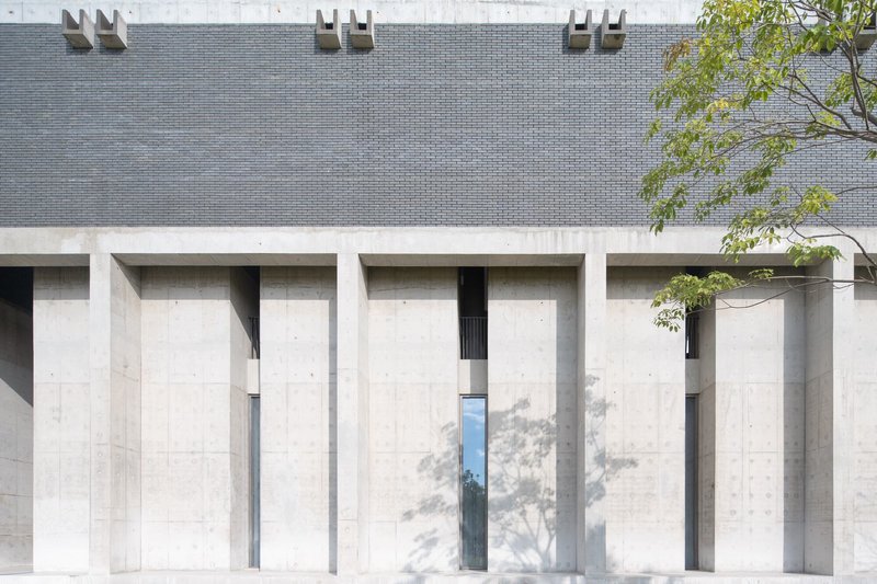 Facade with recessed concrete columns below grey brick wall and dappled tree shadows in daylight