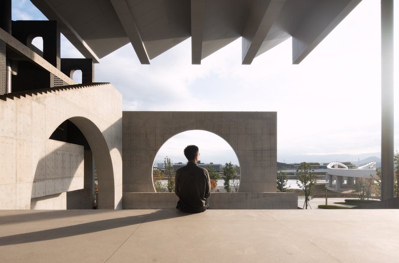Visitor seated on a concrete bench beneath a coffered canopy looking through a circular opening