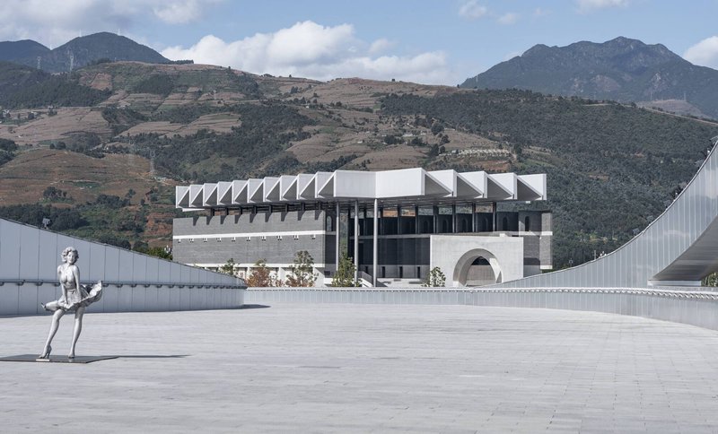 Plaza view with sculptural figure and sawtooth-roofed volumes set against rugged mountain terrain