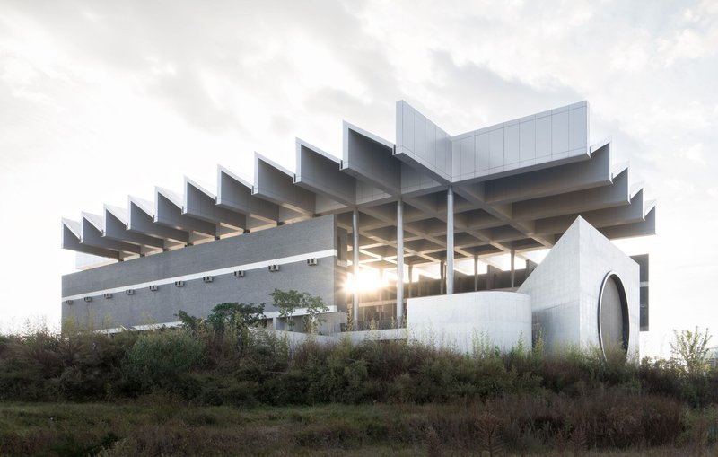 Sawtooth concrete roof structure with exposed columns backlit by sunset glow through vegetation