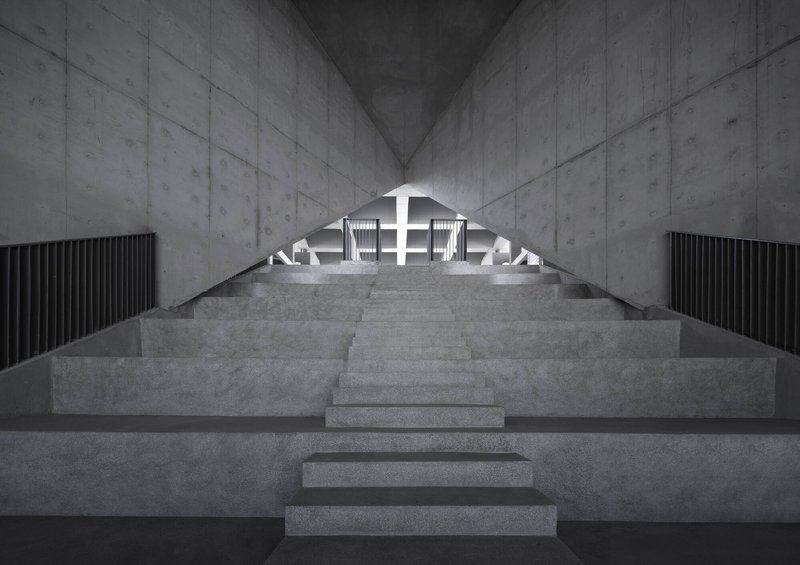 Central concrete staircase rising between board-formed walls toward a backlit arched opening