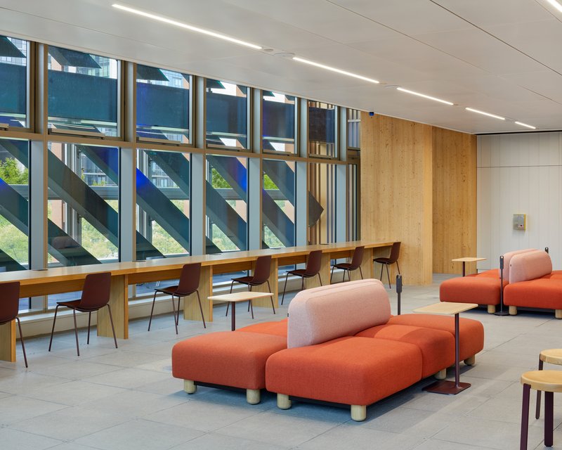 Study area with low orange seating modules and timber desks facing glass curtain wall with view to adjacent building