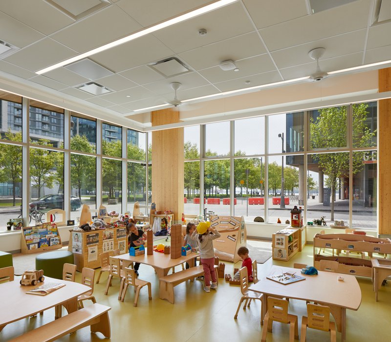 Childcare room with wooden furniture and floor-to-ceiling windows overlooking a tree-lined street