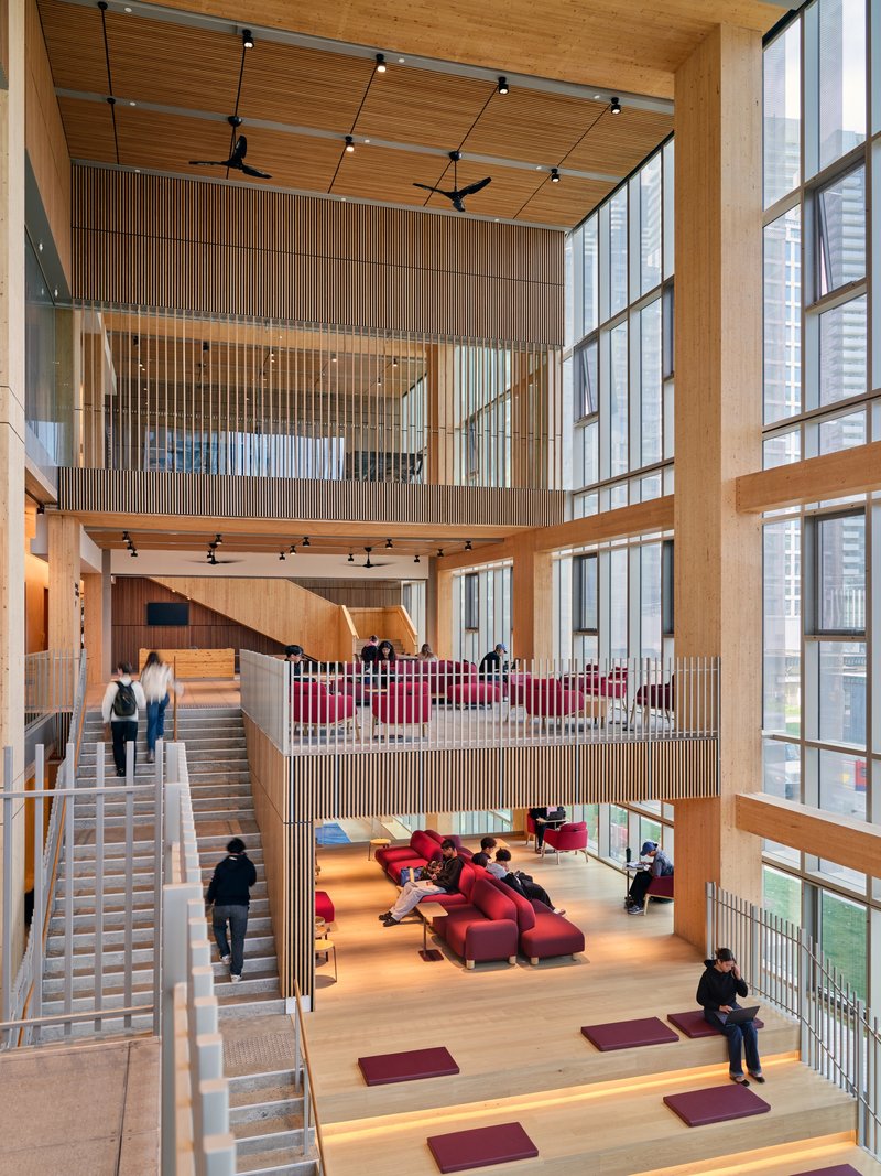 Triple-height atrium with timber screens, stepped platforms, and people gathering on red carpet squares