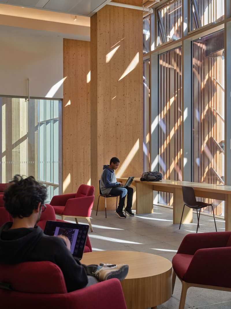 Upper-level lounge with floor-to-ceiling windows casting diagonal shadows across timber columns and study furniture