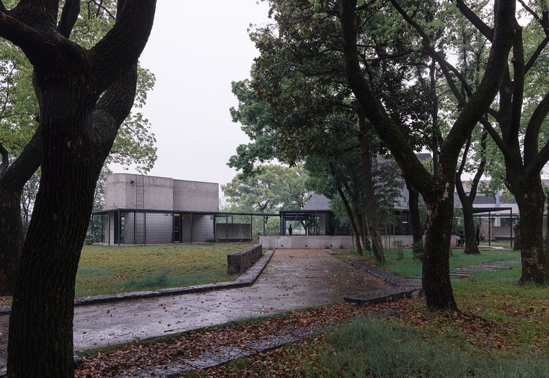 Paved path through mature trees leading to a concrete volume and glass-walled pavilions under overcast skies