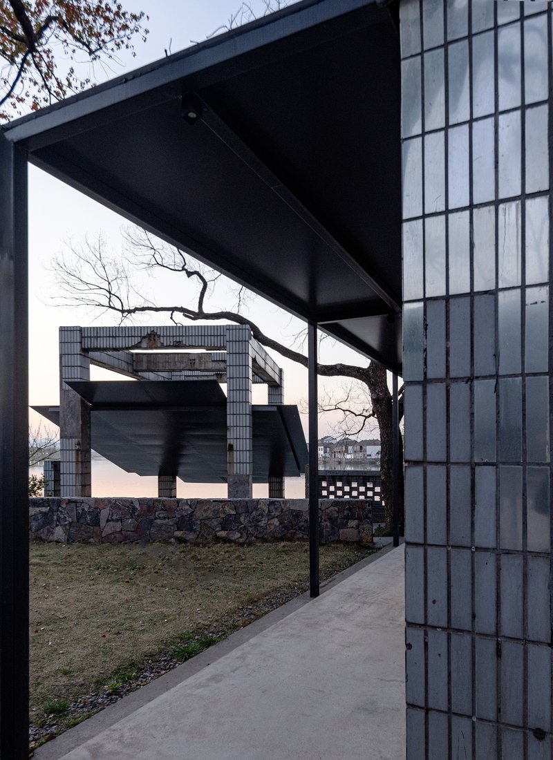 Concrete walkway beneath steel canopy framed by glass block wall and bare tree branches at dusk