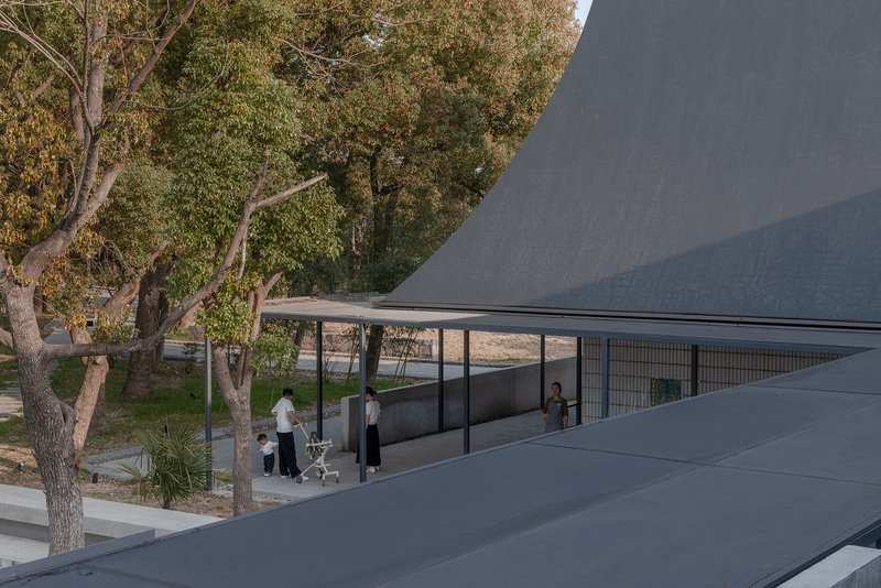 Curved gray roofscape framing a glass pavilion sheltered beneath mature trees with visitors below