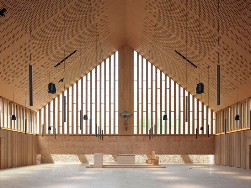 Altar wall with vertical timber screens, crucifix and suspended lighting beneath a pitched wood ceiling