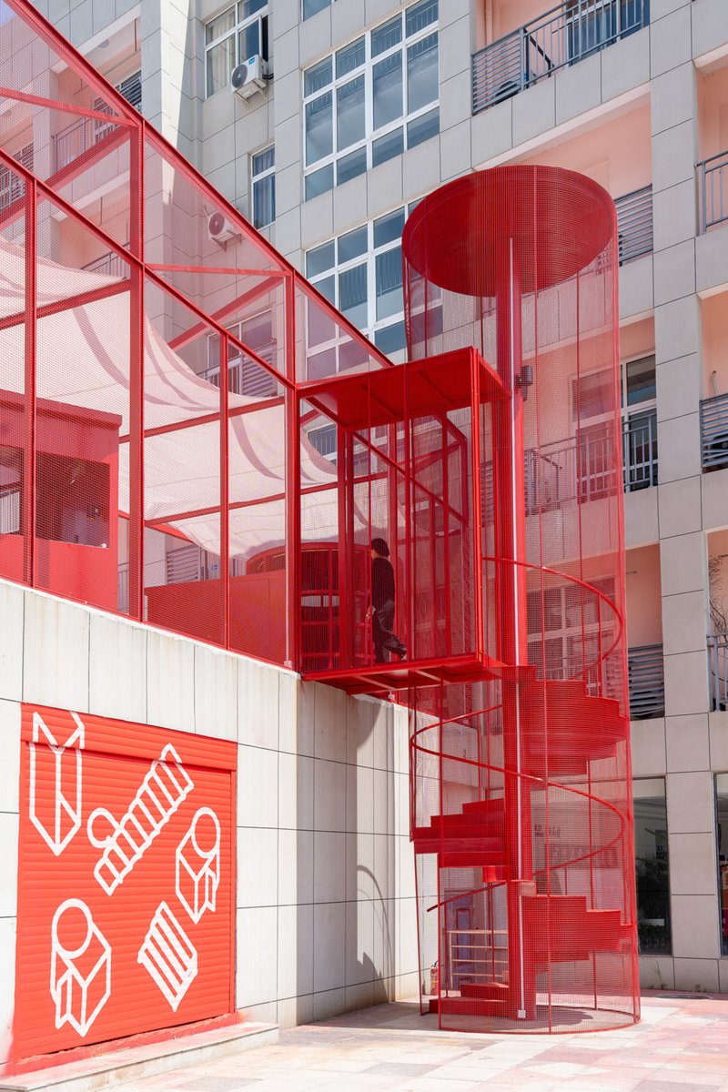 Exterior red spiral staircase enclosed in perforated metal mesh climbing the tiled facade