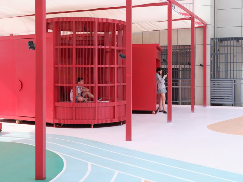 Covered courtyard showing cylindrical red storage pavilion with vertical slats beneath steel frame
