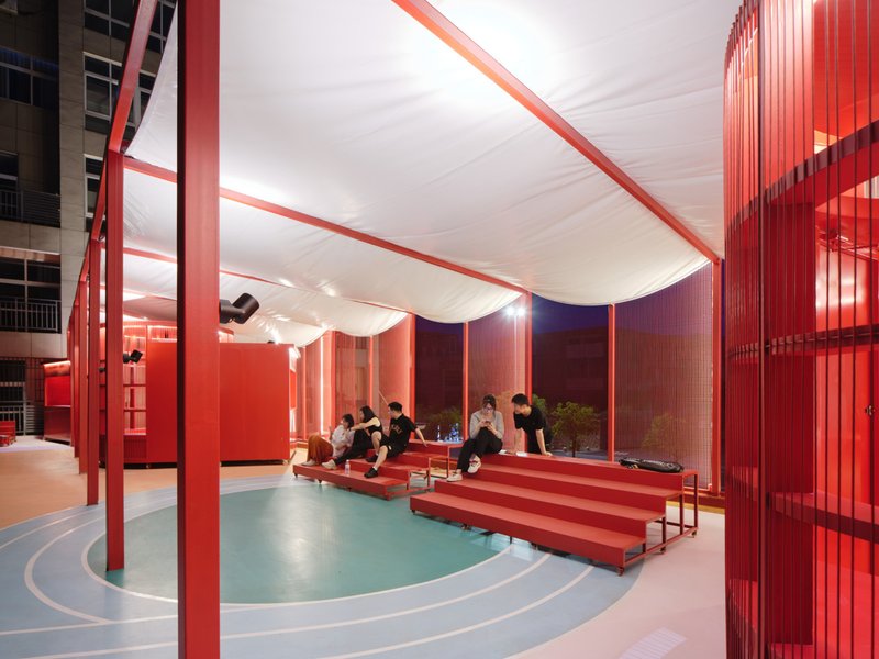 Covered pavilion with red steel columns, white fabric canopy and people seated on stepped benches at dusk