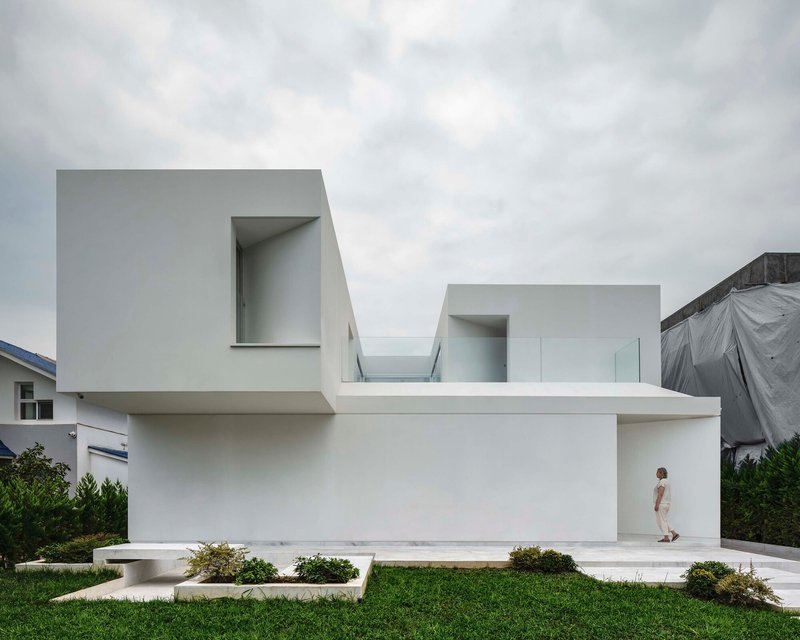White rendered facade with cantilevered volumes and recessed entrance where a figure stands under overcast skies
