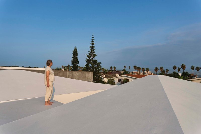 Rooftop terrace with white rendered volumes and a figure overlooking palm trees in the distance