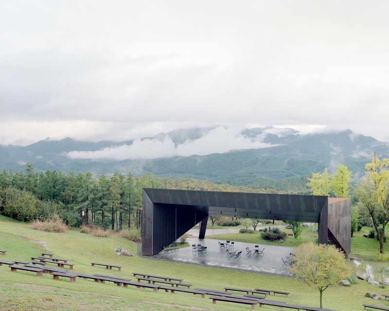 Cantilevered steel volume sheltering an outdoor seating terrace with grassy amphitheater and misty mountains beyond