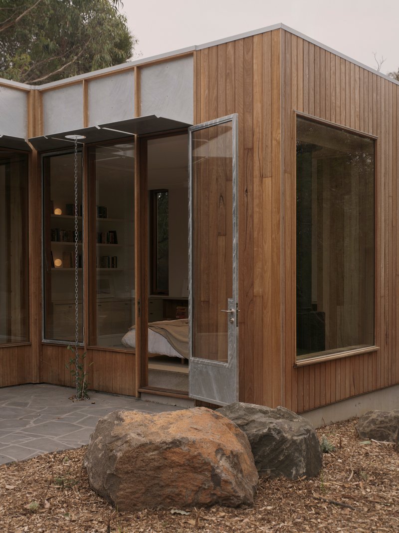 Timber-clad bedroom pavilion with glazed door and corner window beside boulders on mulched ground