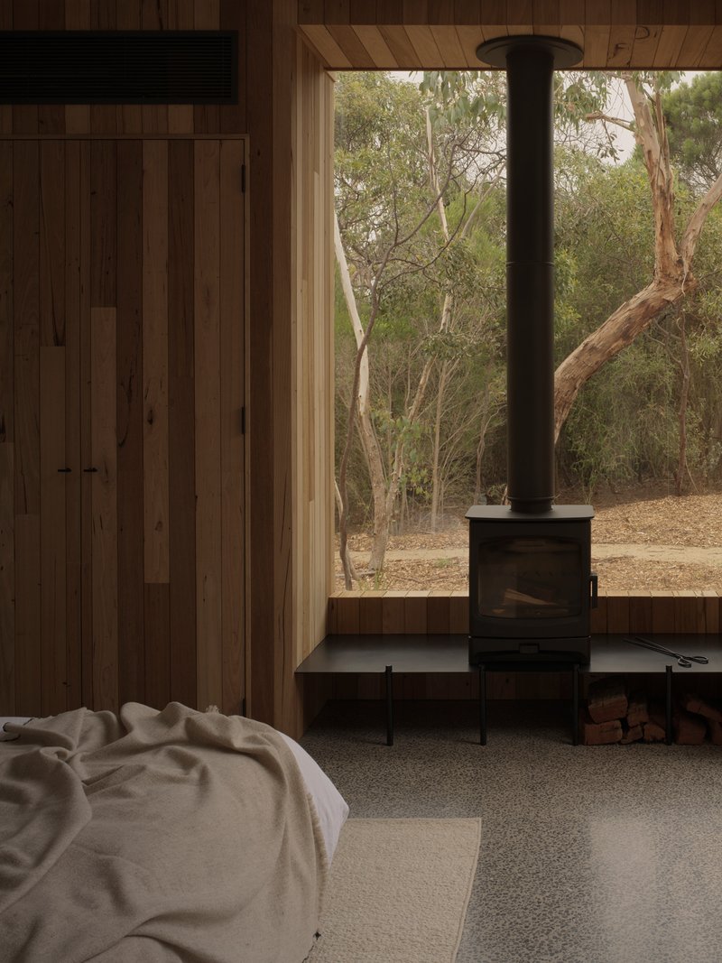 Interior view of freestanding black wood stove on steel plinth with timber wall and window overlooking trees