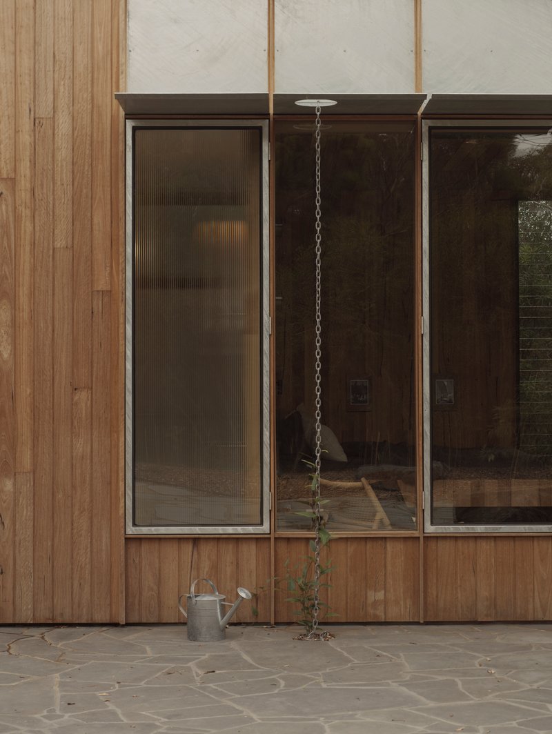 Timber-clad facade with tall glazed panels above a stone terrace with hanging plant and watering can