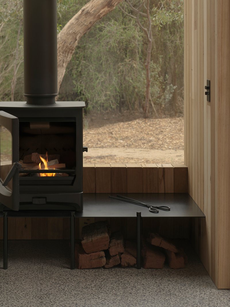 Black freestanding wood stove with integrated firewood storage beside a glazed door opening to trees