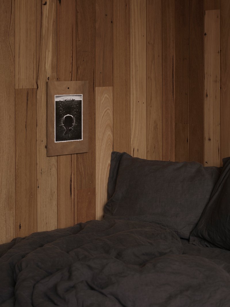 Framed photograph mounted on vertical timber wall above dark grey bed linens in dim light