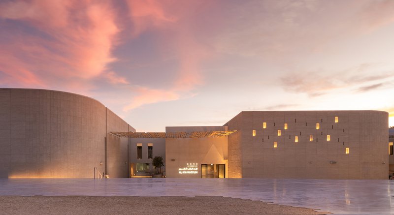 Plaza view at sunset showing perforated stone volumes and a timber pergola canopy under pink clouds