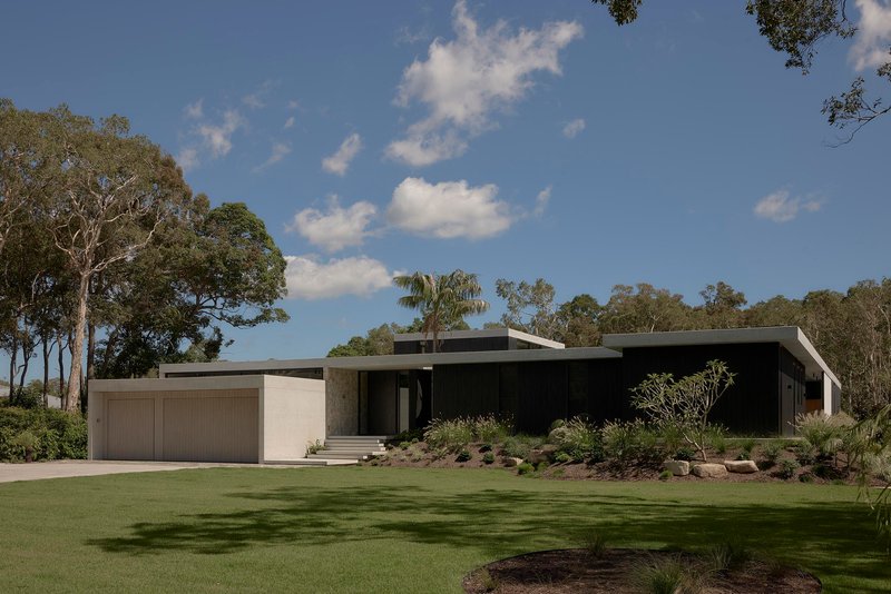 Long horizontal facade with black cladding and sandstone garage in a lawn clearing surrounded by eucalyptus trees