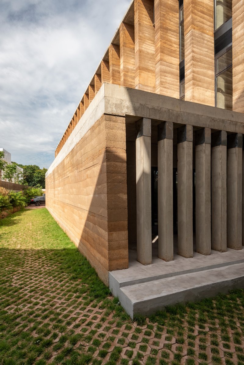 Corner detail showing the concrete beam cantilevering over cylindrical grey columns and rammed earth wall