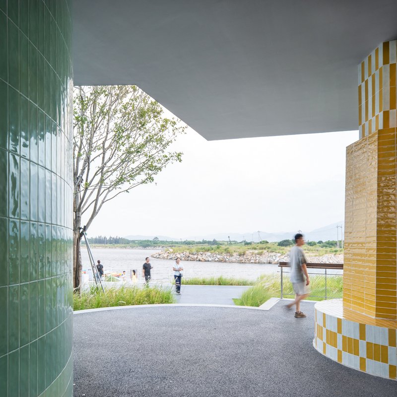 Covered walkway framed by green tiled wall and yellow mosaic column opening to waterfront grasses
