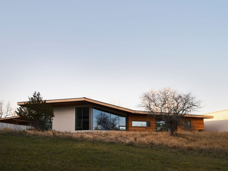 Long timber roof cantilever extending over white stucco and wood-clad volumes in dry grassland at dusk