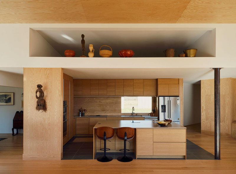 Open kitchen with light wood cabinetry, island with two stools, and recessed display shelf above