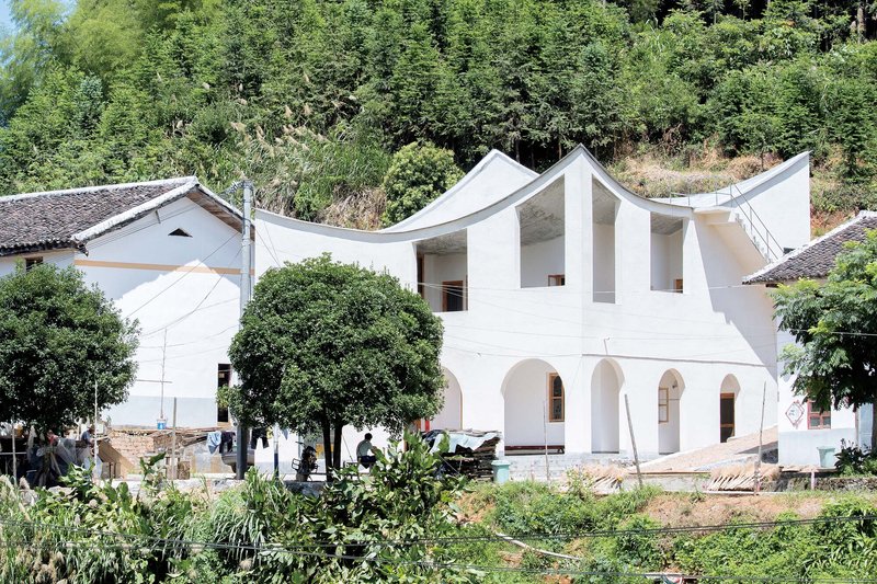 White facade with arched openings and three undulating roof peaks framed by lush vegetation and neighboring houses
