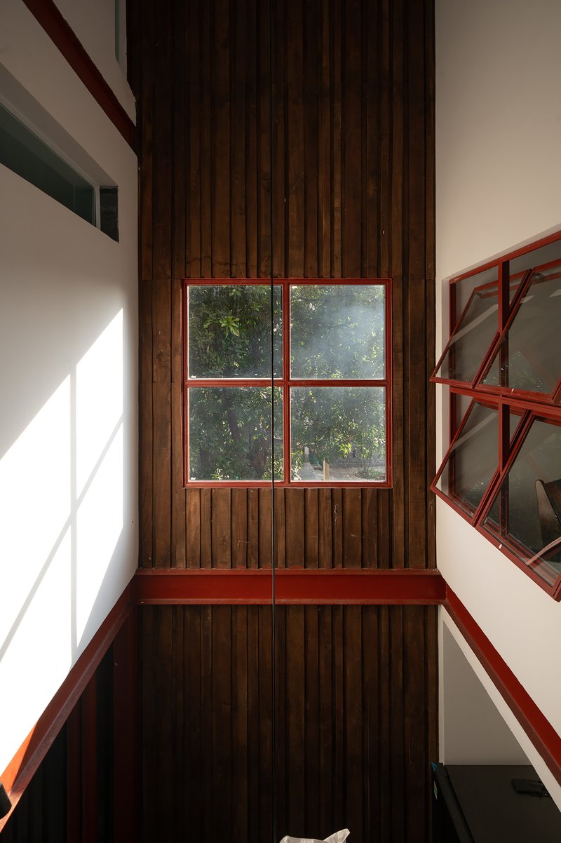 Double-height stairwell with red steel framed window set in vertical timber cladding under strong sunlight