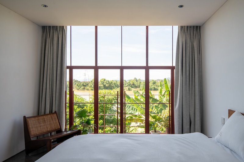 Bedroom interior with floor-to-ceiling red steel framed windows overlooking banana palms and river